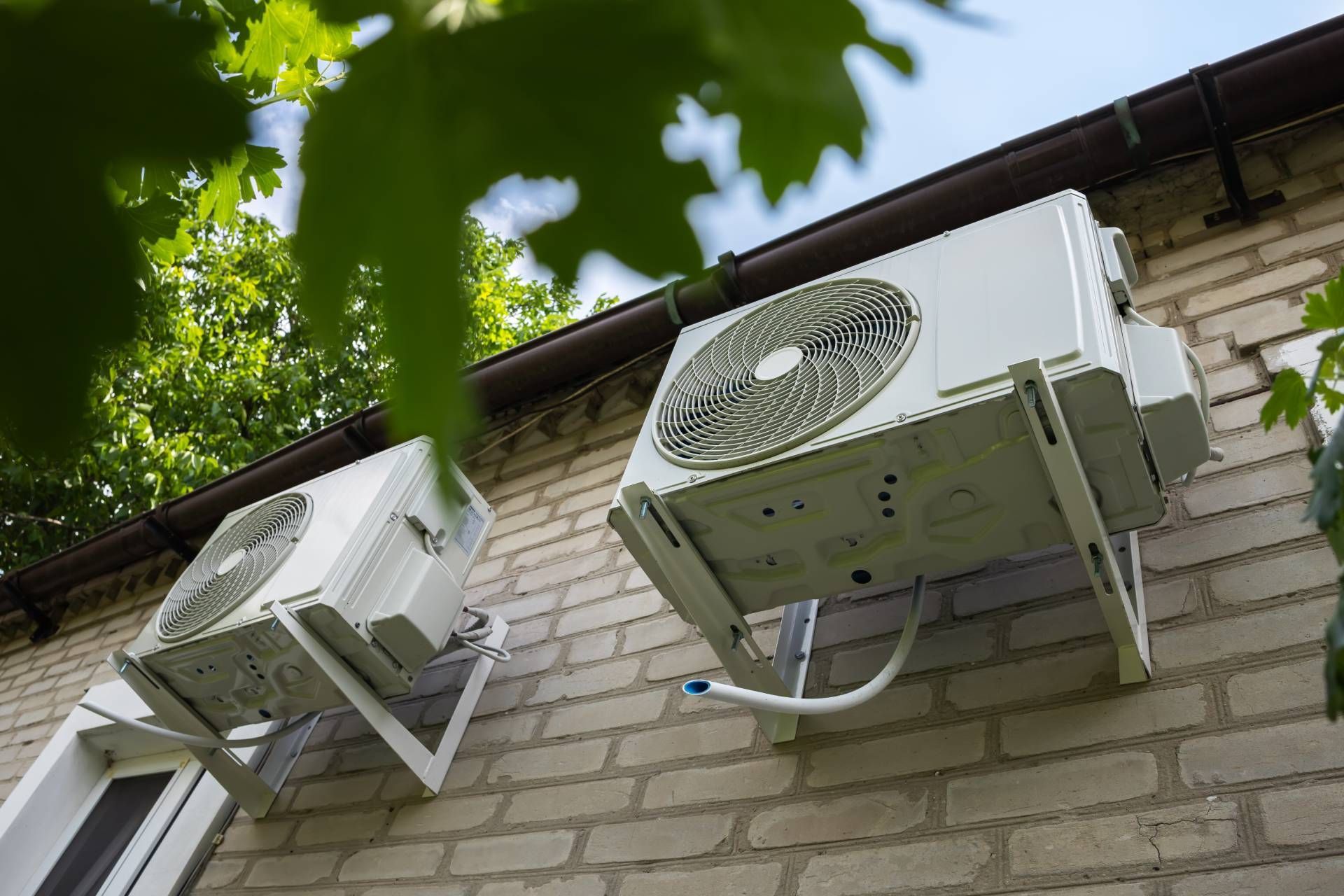 Two white outdoor air conditioner units mounted on a brick wall beneath green leaves.