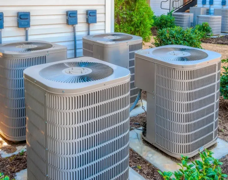 Four outdoor air conditioning units beside a building on a paved area with greenery nearby