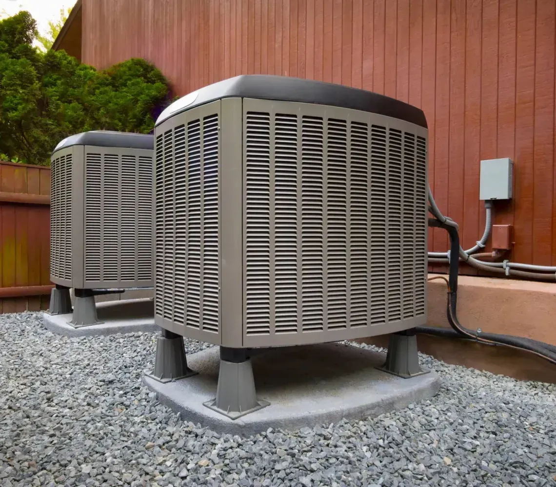 Two outdoor HVAC units on gravel beside a wooden building, with a red fence in the background.