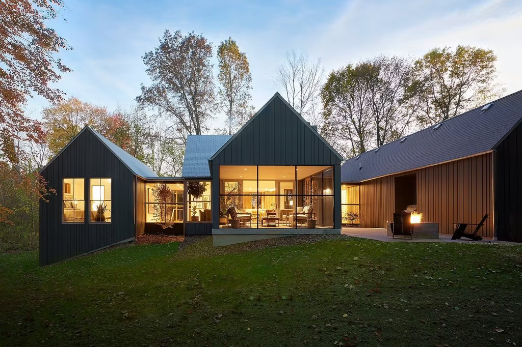 Modern dark-wood house with lit windows and glass walls at dusk, surrounded by trees and lawn