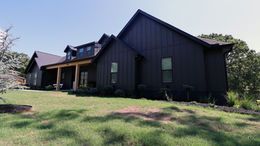 Dark brown house with a covered porch and upper deck, viewed from a grassy yard under a sunny sky
