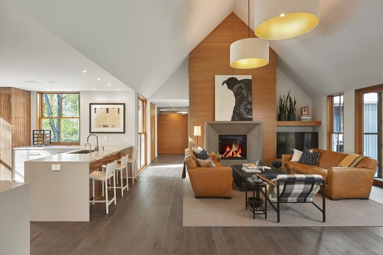 Modern open-concept living room with vaulted ceiling, fireplace, tan sofa, and adjacent white kitchen