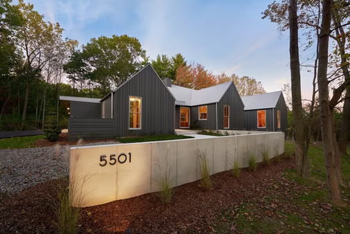 Modern gray house with lit windows behind a low wall marked 5501, surrounded by trees at dusk