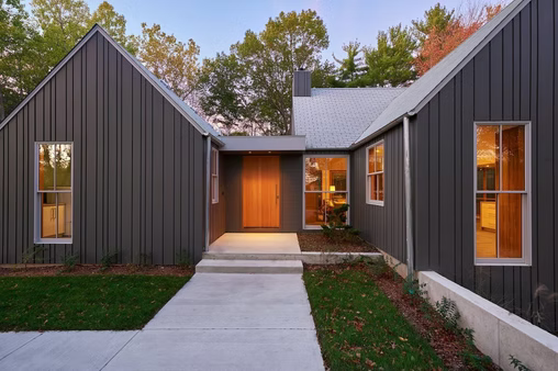 Modern dark-gray house with gabled roofs, lit entryway, and concrete walkway at dusk