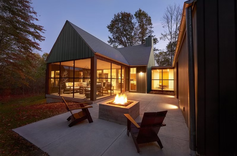 Modern glass house patio at dusk with a fire pit and two lounge chairs