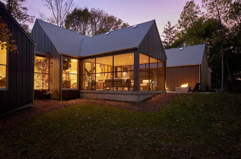 Modern glass-walled house glowing at dusk with warm interior lights and a dark metal roof