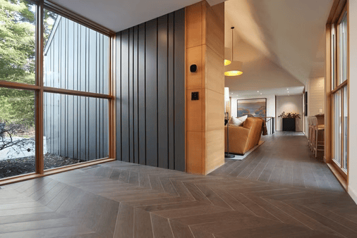 Modern wood-paneled entry hall with herringbone floor, glass wall, and view into a bright living room