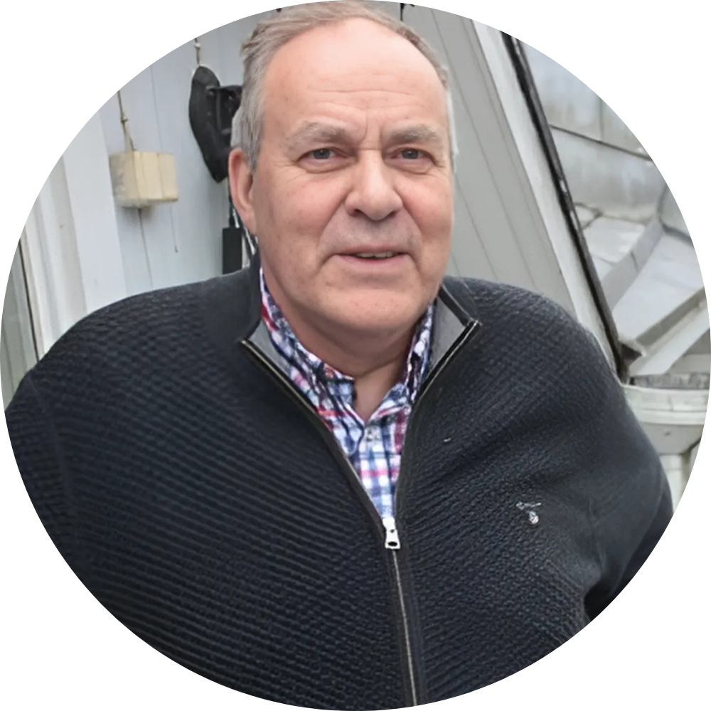 Man in black sweater, plaid shirt, smiling outdoors.