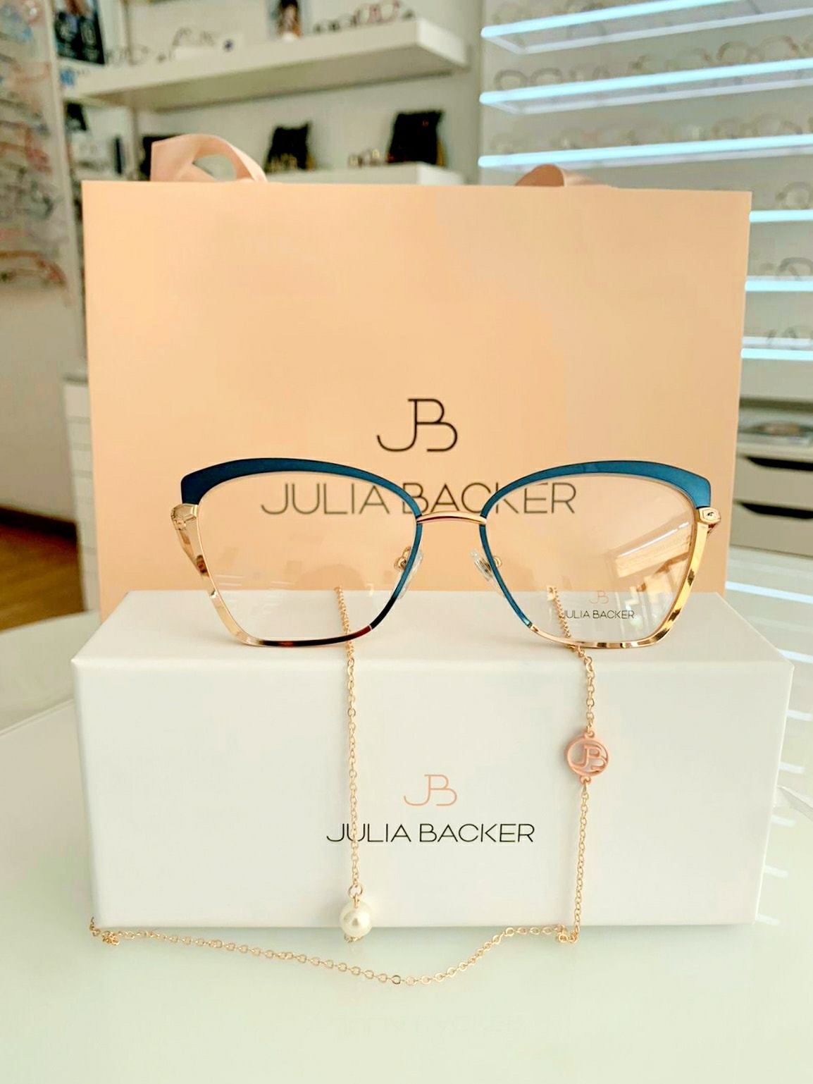 Occhiali da vista Julia Backer con montatura color verde acqua e oro e catena decorativa, appoggiati su una scatola con il marchio in un negozio di ottica.