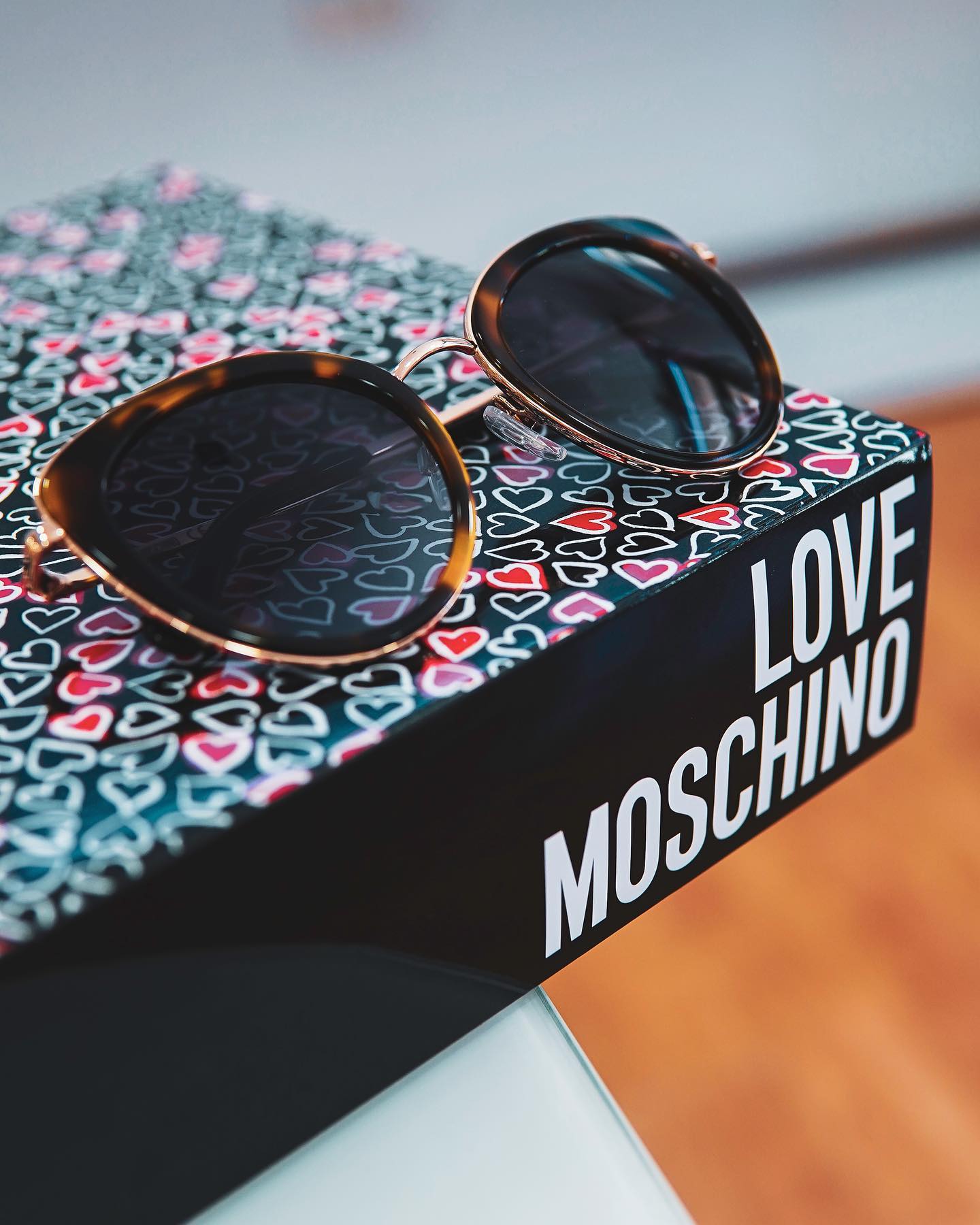 Gli occhiali da sole cat-eye in tartaruga poggiano su una scatola nera decorata con piccoli cuori e la scritta bianca LOVE MOSCHINO.