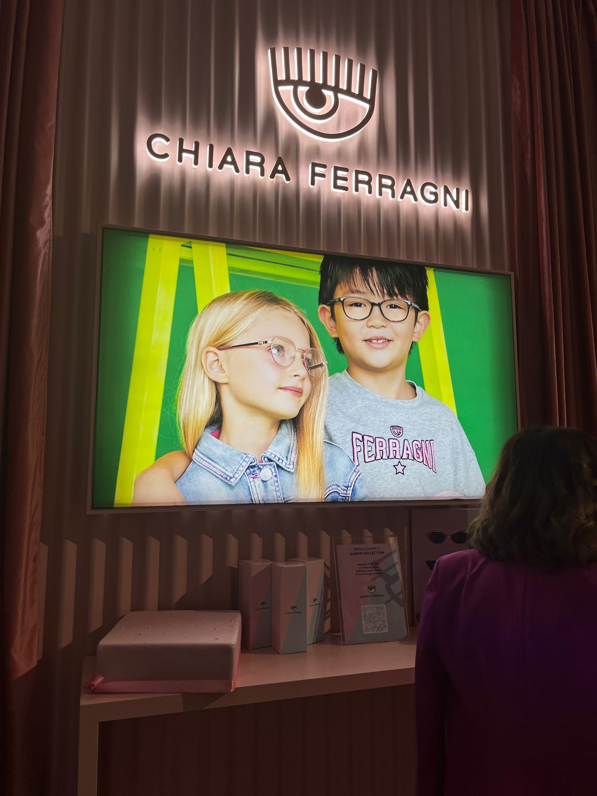 Uno schermo che mostra due bambini che indossano occhiali sotto il logo luminoso di Chiara Ferragni in una boutique.