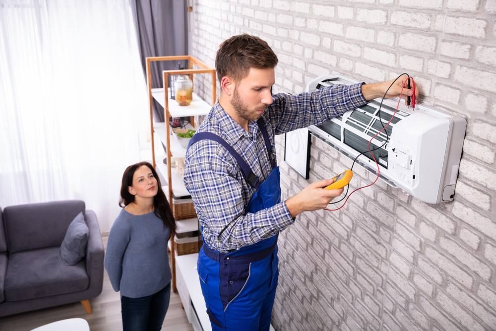 HVAC technician uses multimeter on a wall-mounted AC unit; a woman watches in a room.