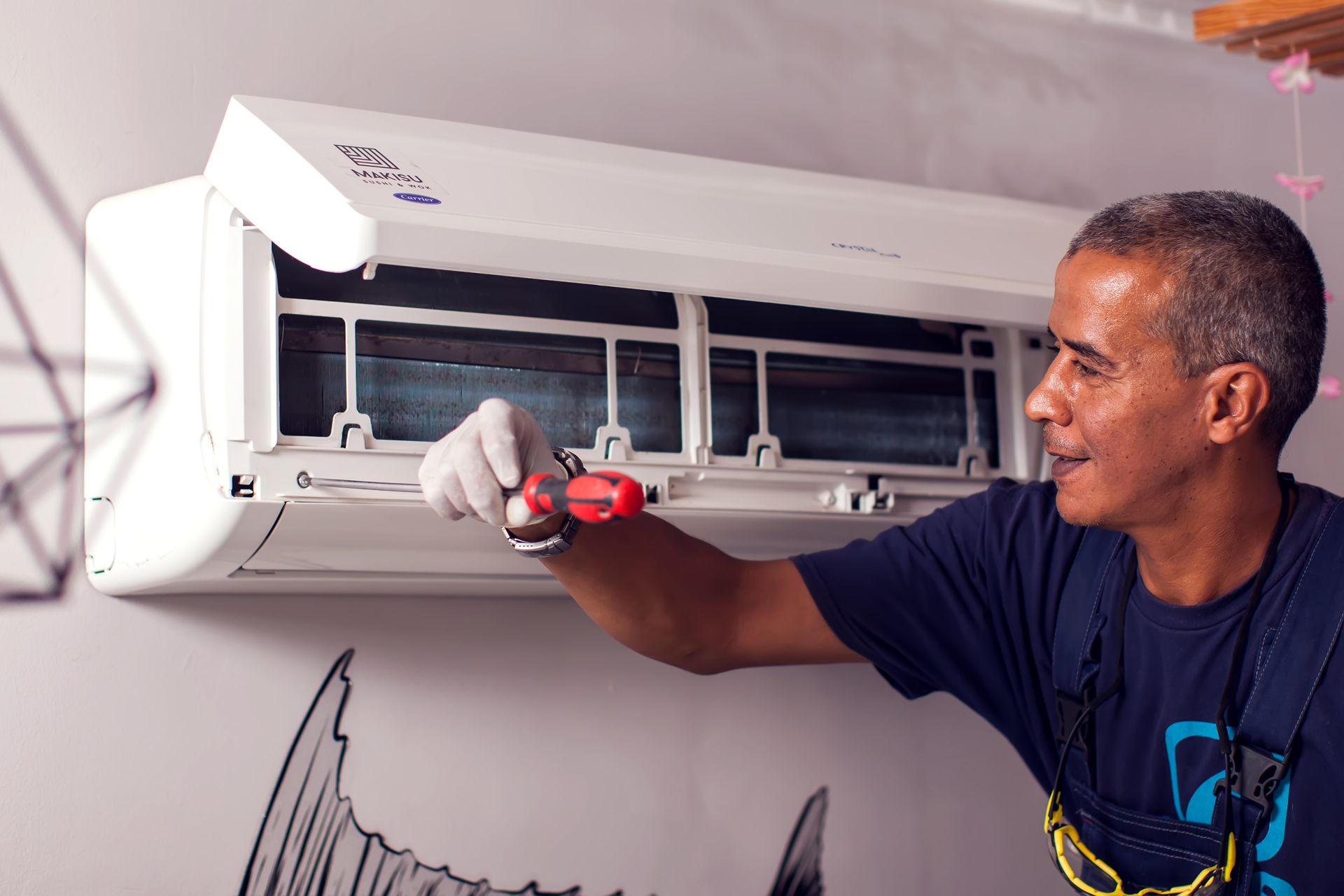 Man in work overalls inspects an air conditioner with a screwdriver. White unit on wall, in a room.