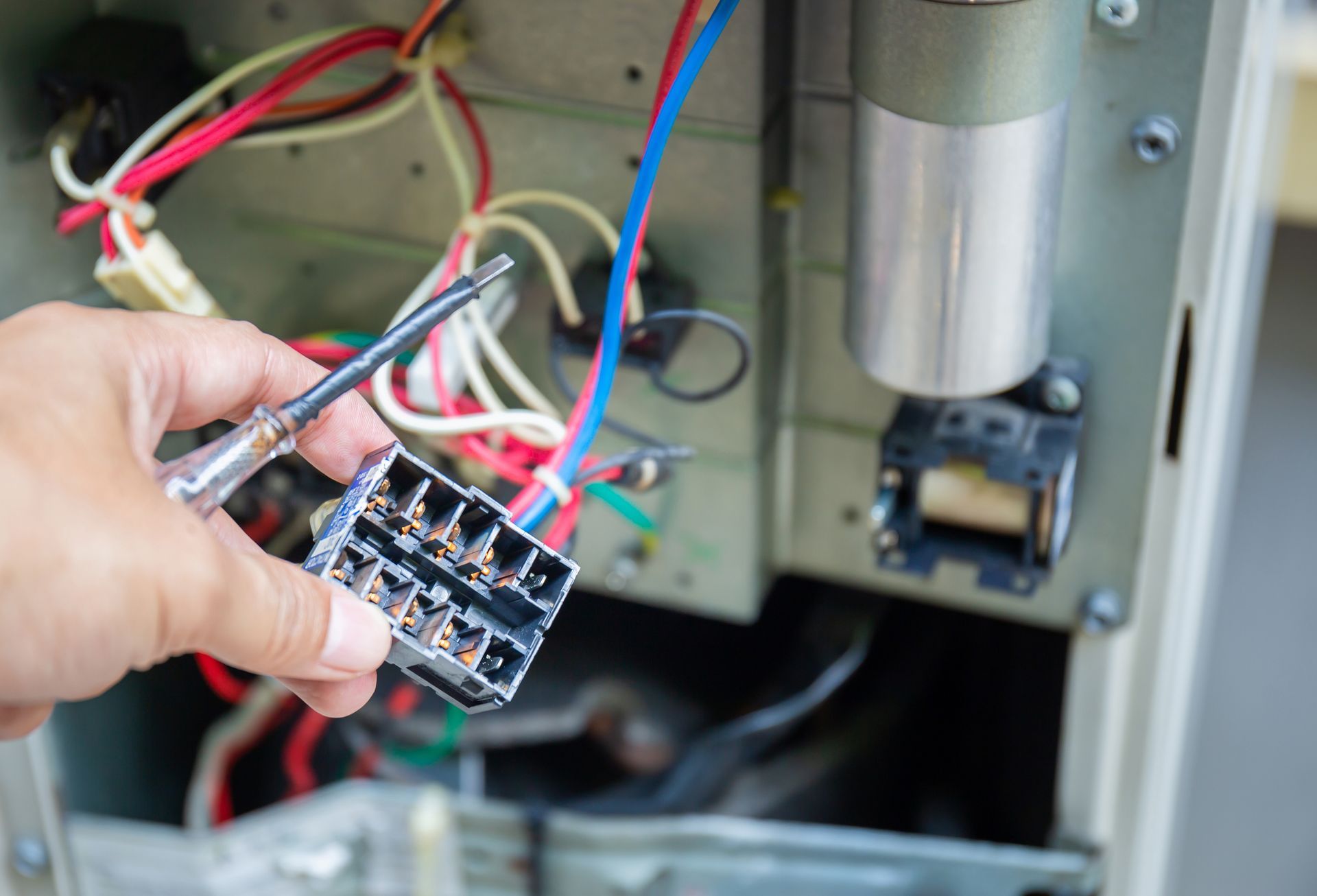 Hand holding a screwdriver near exposed electrical wiring and a connector within a machine cabinet.