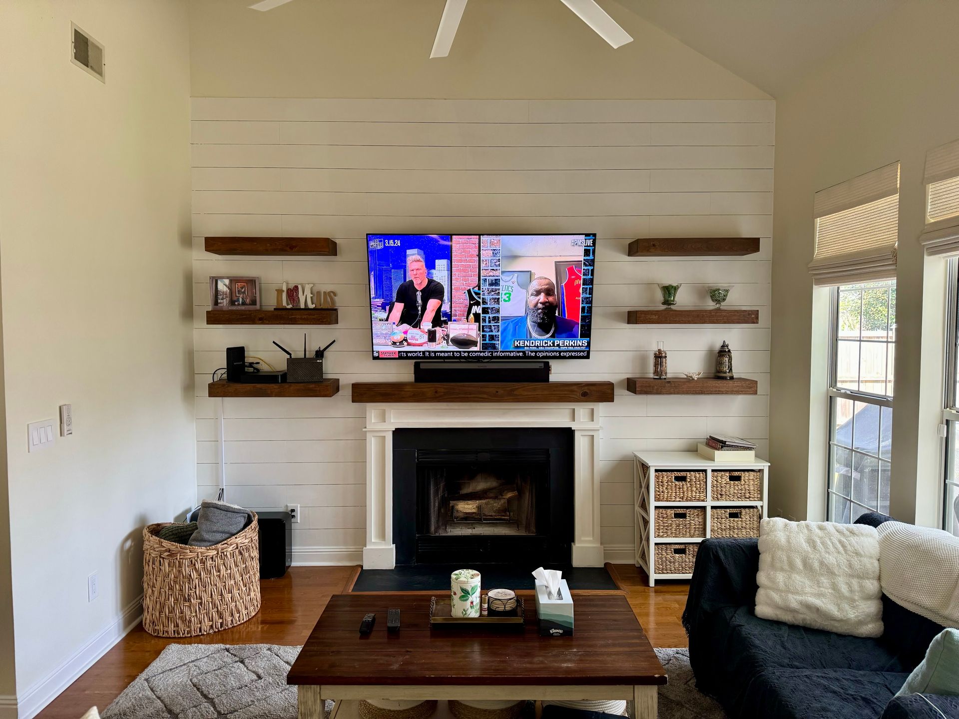 Bright living room with white fireplace, wall-mounted TV, shelves, and dark sofa by a window