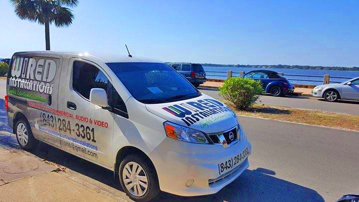 White branded van parked by a waterfront road under a blue sky, with palm trees and cars in the background.