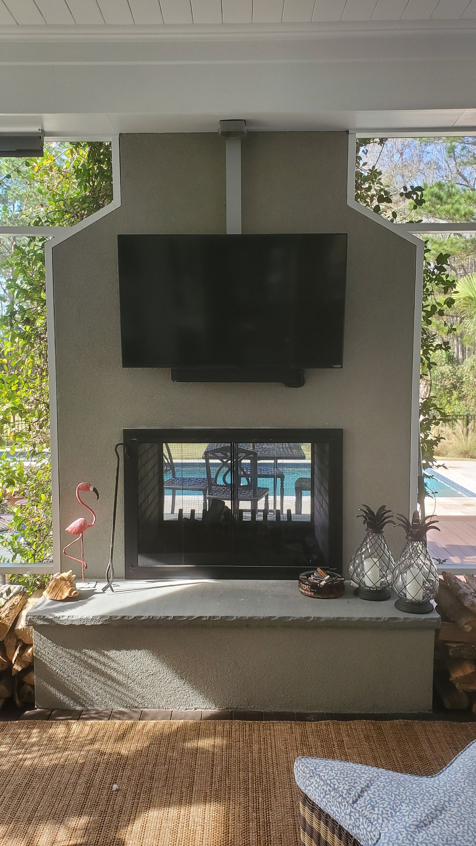 Patio with wall-mounted TV above a fireplace, framed by open side views and a wicker chair in the foreground