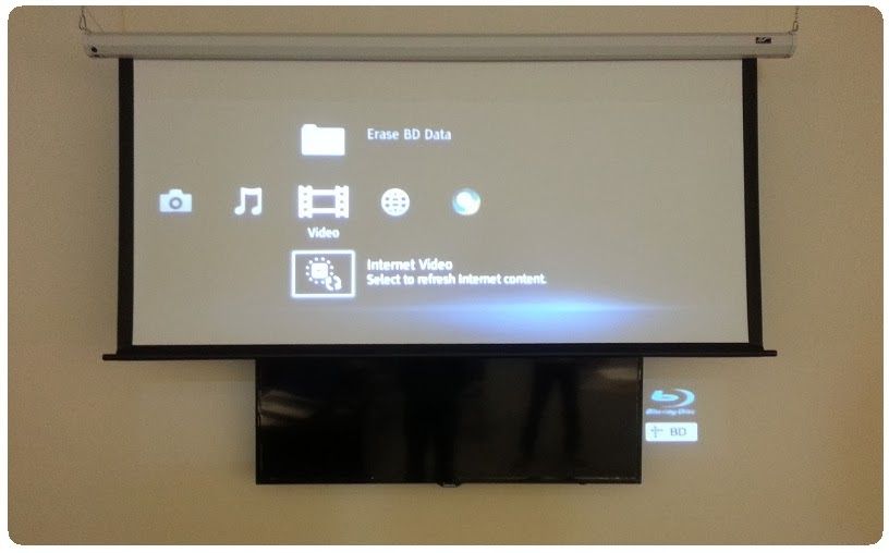 Projector screen showing a media player menu above a wall-mounted TV in a room