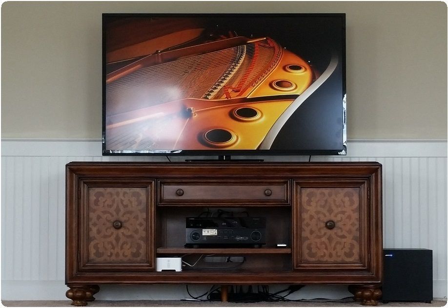 Flat-screen TV above an ornate wooden cabinet in a beige room, with a small black speaker on the floor.