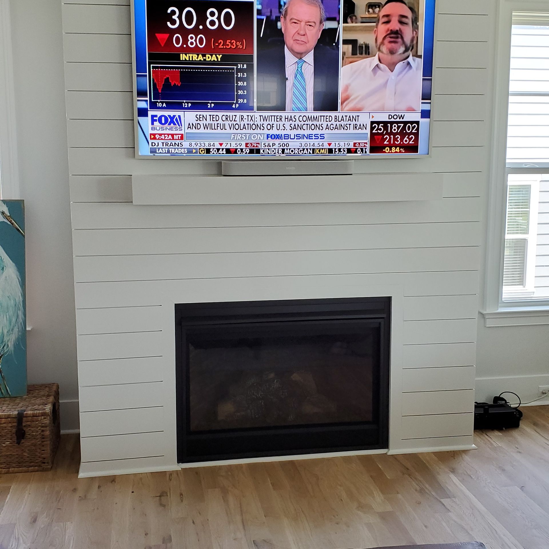 Living room with a white fireplace, wall-mounted TV showing a news broadcast, and light wood floors.