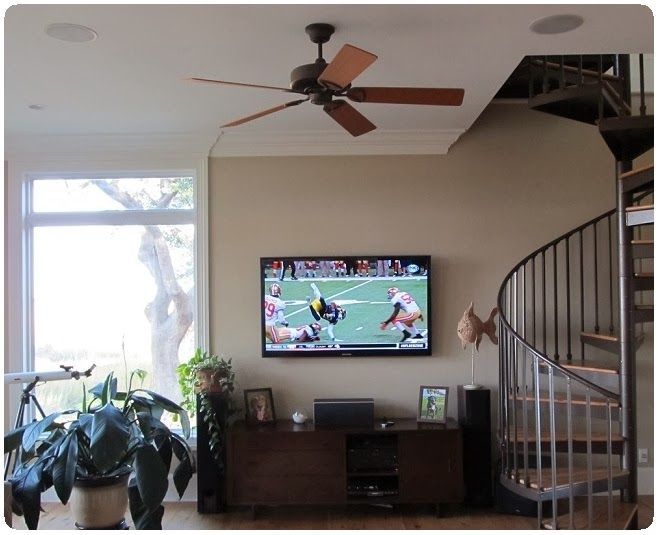 Living room with wall-mounted TV showing football, ceiling fan, potted plants, and spiral staircase