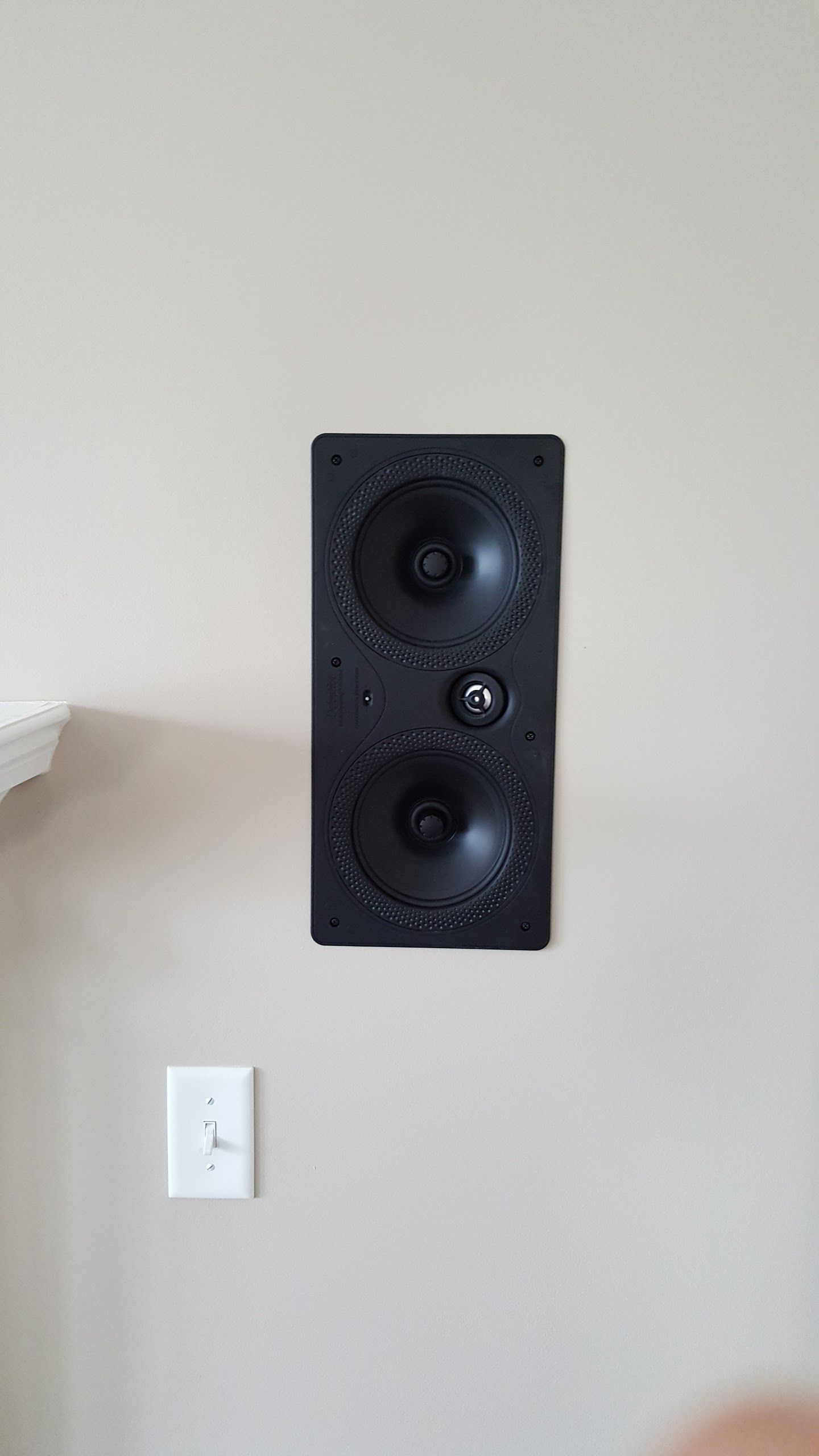 Black wall-mounted speaker on a beige wall above a white light switch.