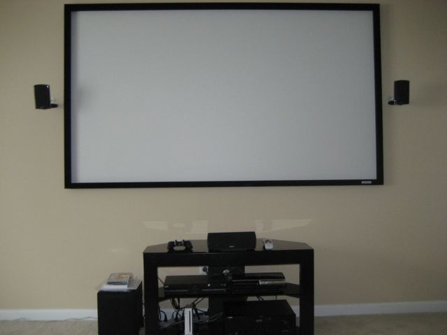 Living room wall with a large projection screen above a black media stand and speakers