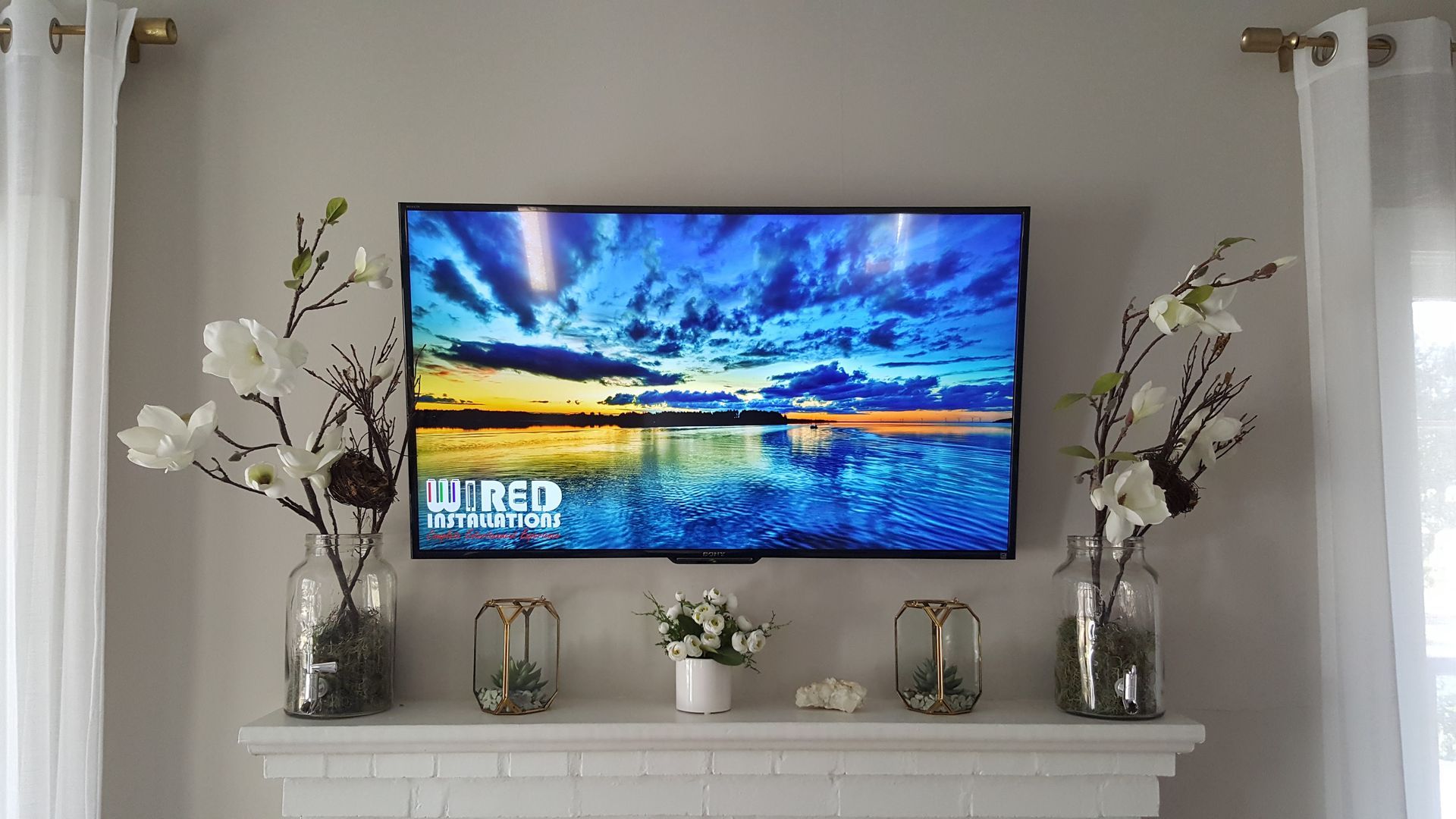 Wall-mounted TV above a white mantel with silver vases, white flowers, and a bright landscape on screen