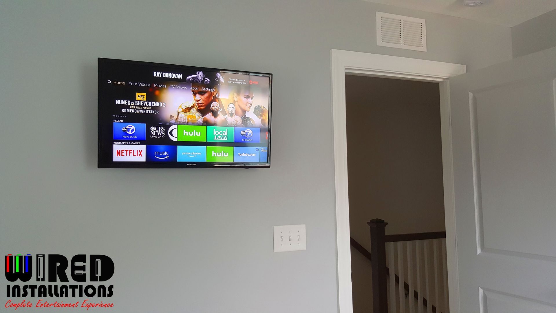 Wall-mounted TV displaying a streaming menu on a gray wall beside an open doorway and staircase.