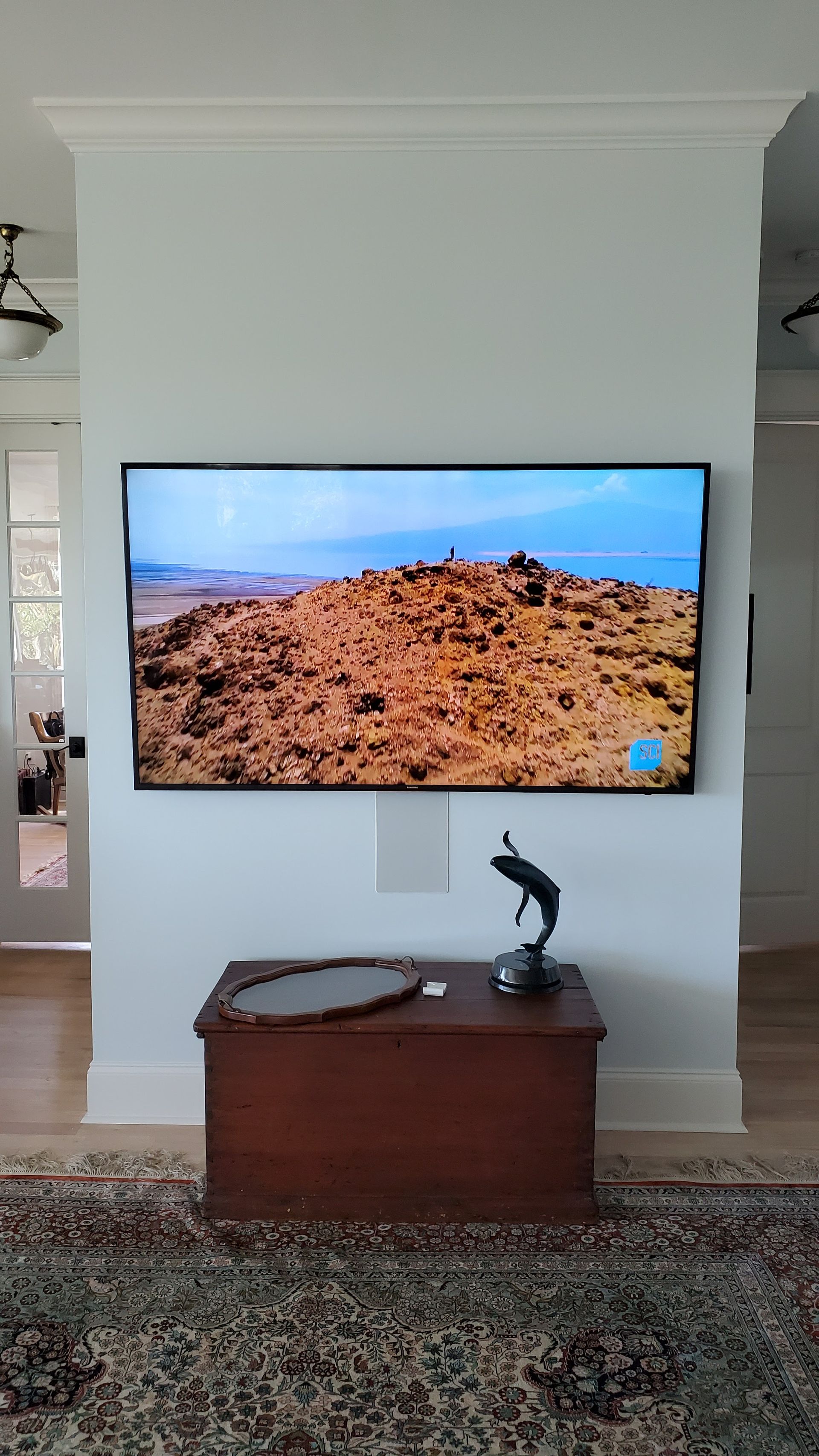 Wall-mounted TV showing a rocky landscape above a wooden cabinet with a sculpture in a bright room