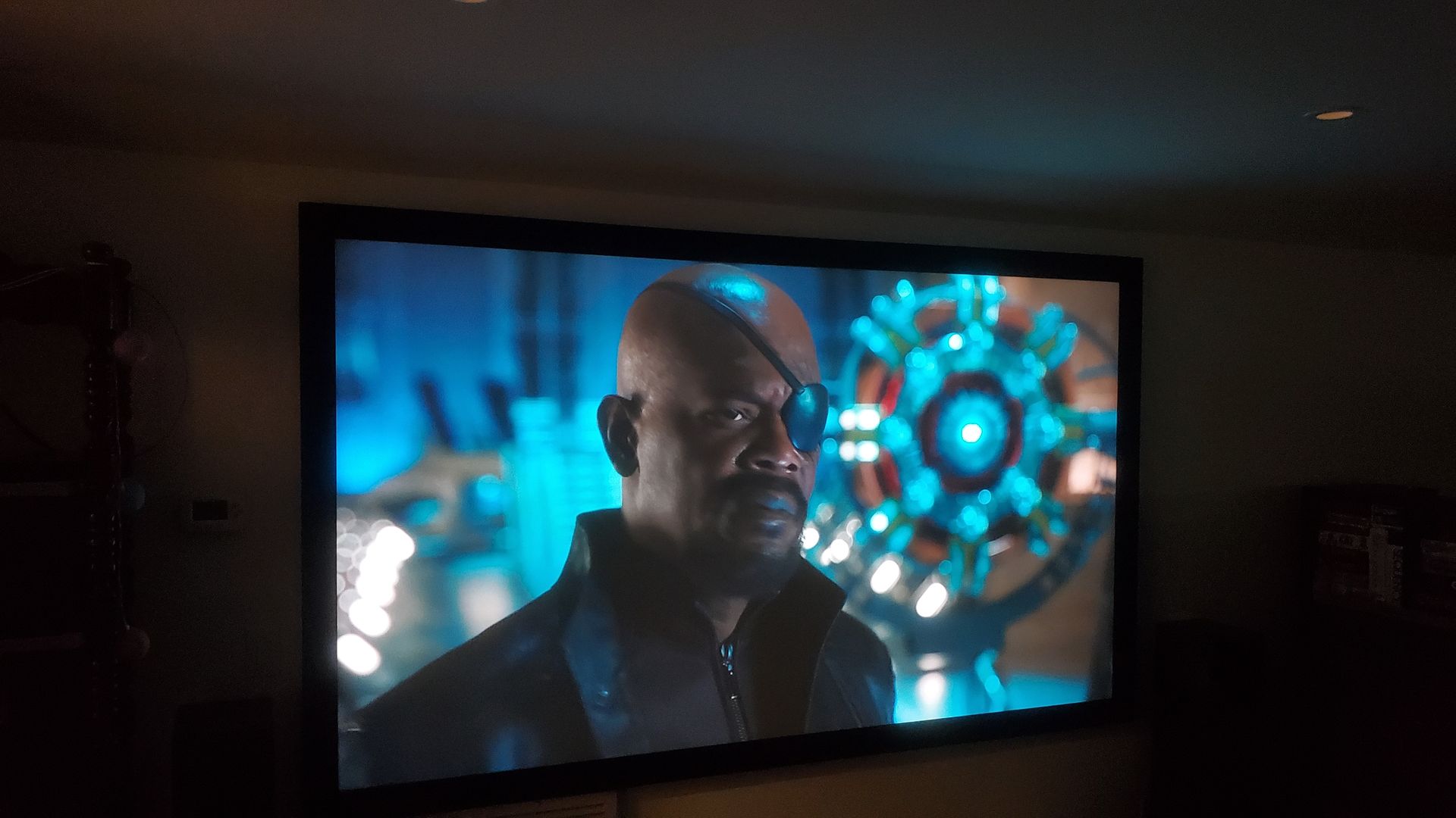 A TV screen shows a serious man in dark clothing with a glowing blue sci-fi device behind him.