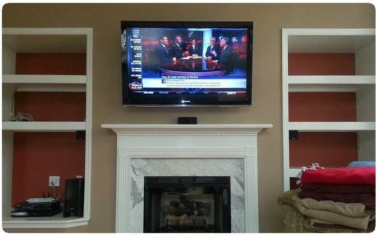 Living room with wall-mounted TV above a fireplace, flanked by built-in shelves and folded blankets on the right.