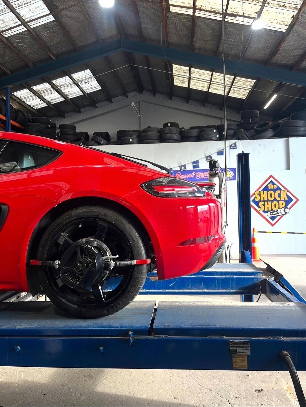 Red sports car on a lift in an auto shop, with wheel removed and a Shock Shop sign nearby