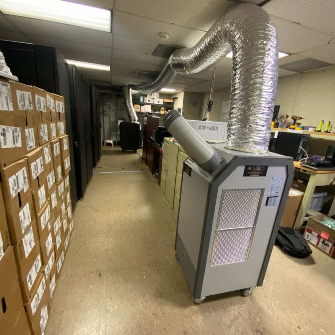 A portable air conditioner unit with a silver exhaust vent hose in a room with stacks of cardboard boxes.