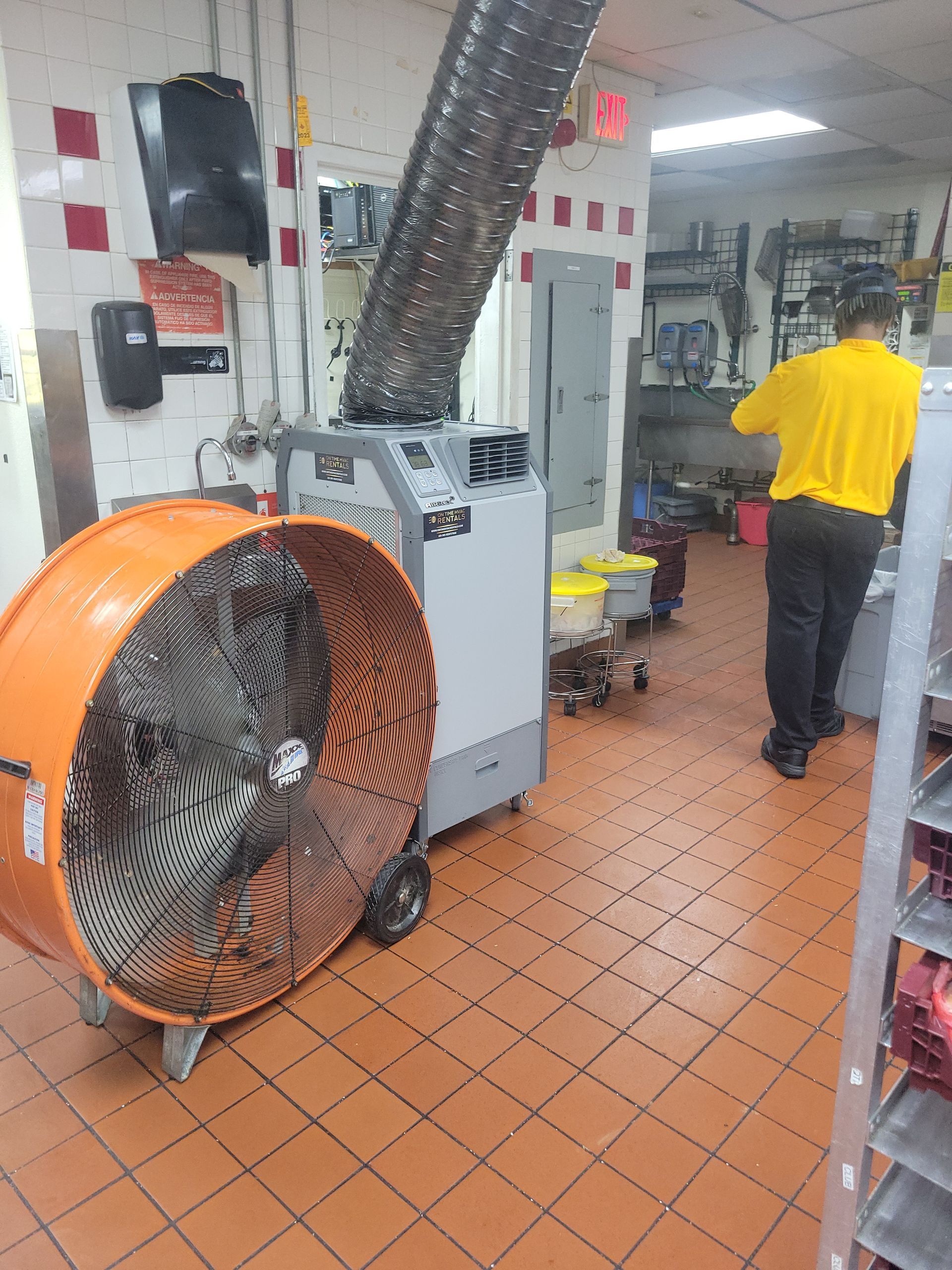 A worker in a yellow shirt stands in a commercial kitchen next to a large orange industrial fan and a portable AC unit.