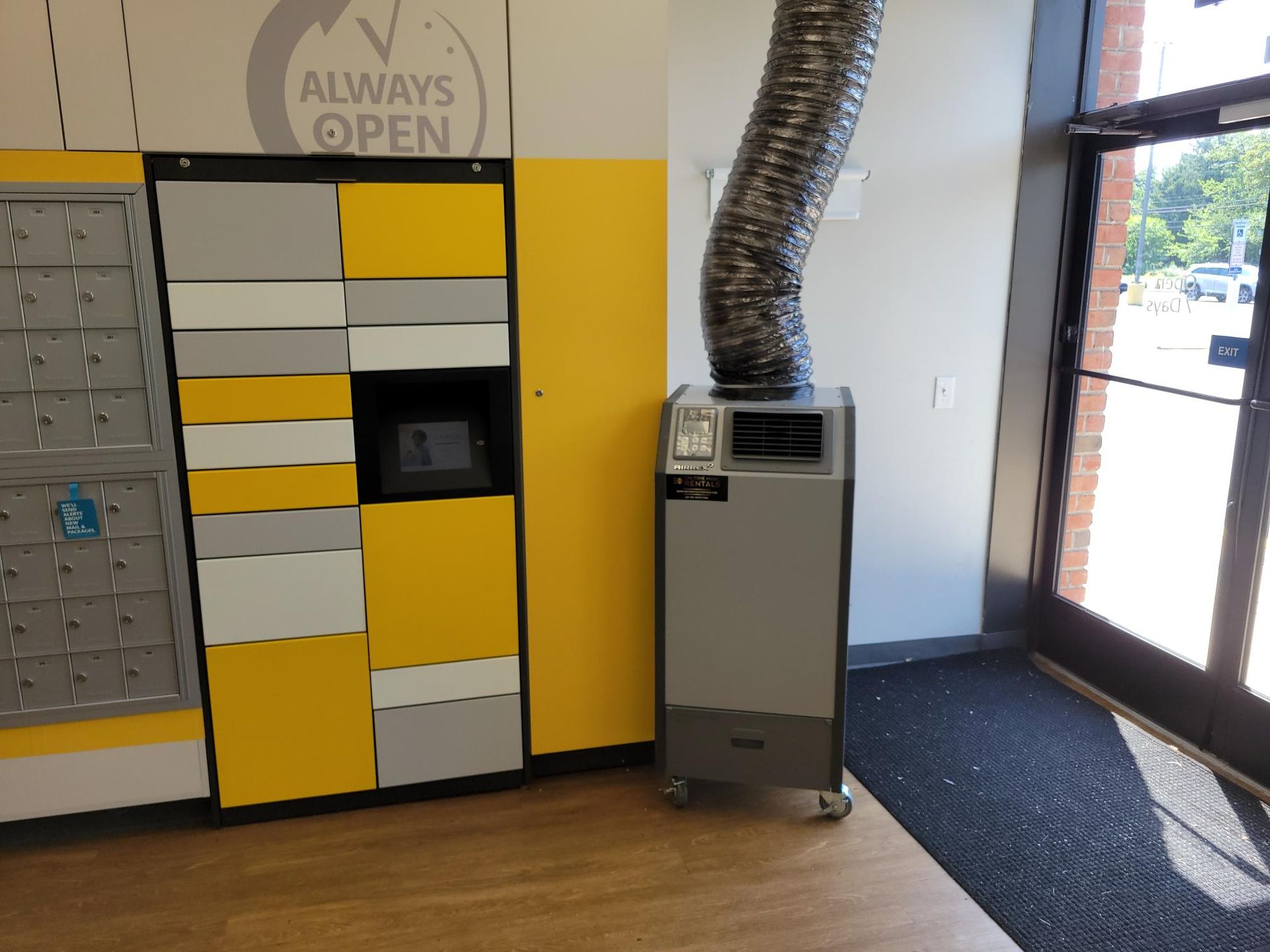A portable air conditioner with a silver exhaust vent stands next to a yellow and gray package locker unit indoors.