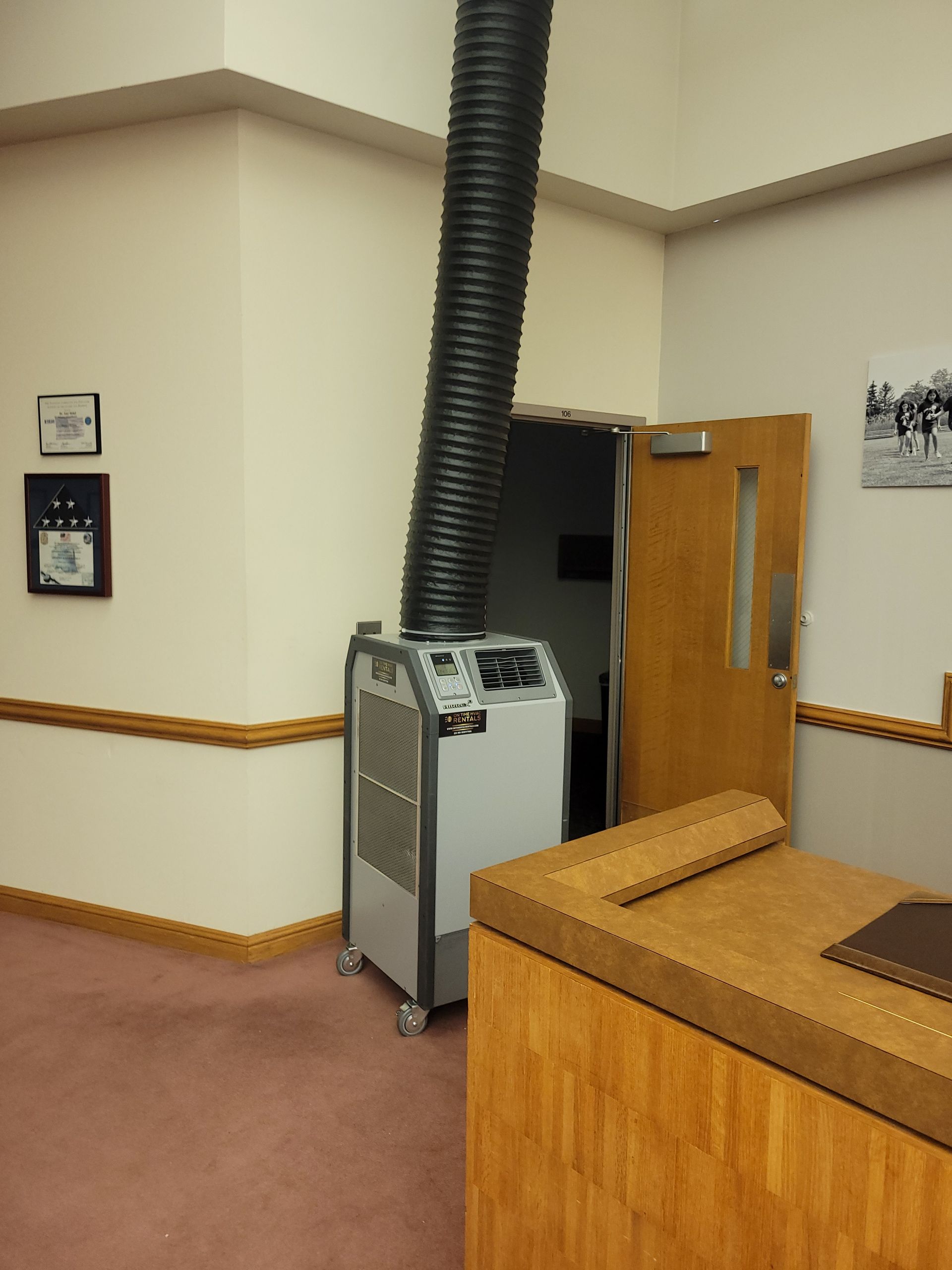 A portable, light-grey industrial air conditioning unit with a black exhaust hose stands in an office hallway.