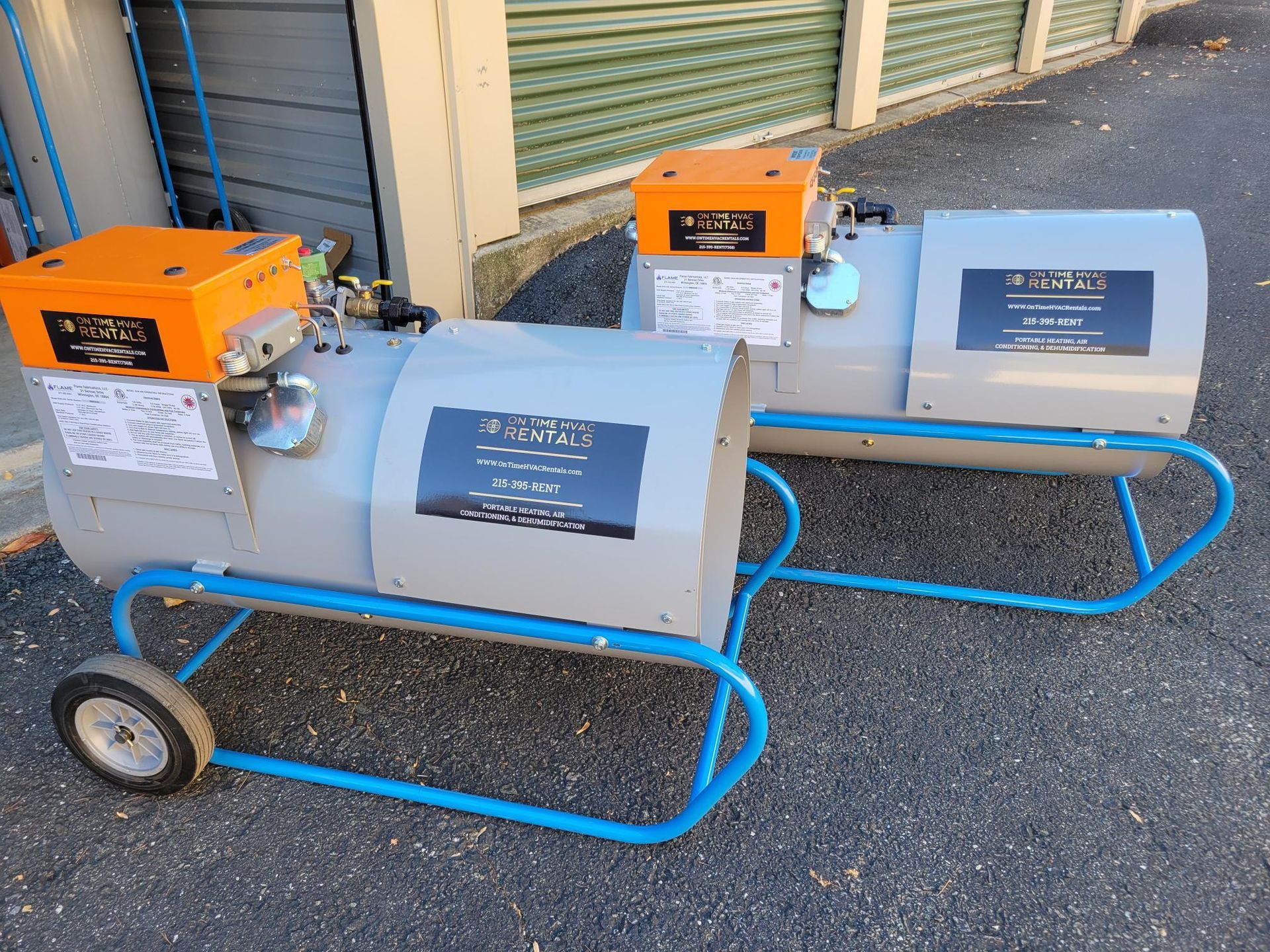 Two portable, grey industrial heaters with orange control boxes on blue wheeled frames sit on asphalt outside a storage unit.
