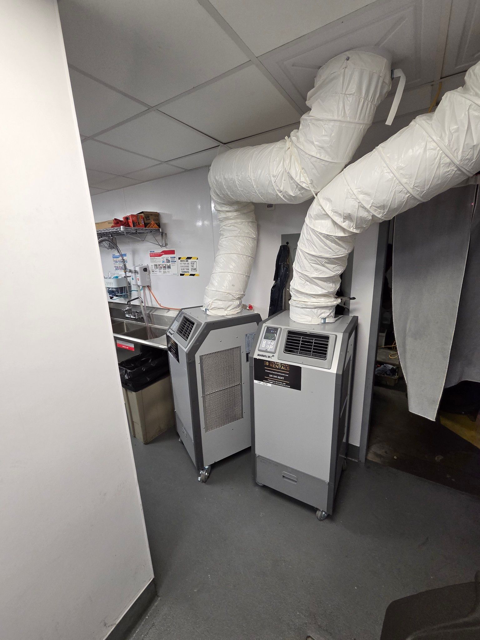 Two portable industrial air conditioners in a room with white exhaust hoses connected to a drop ceiling.