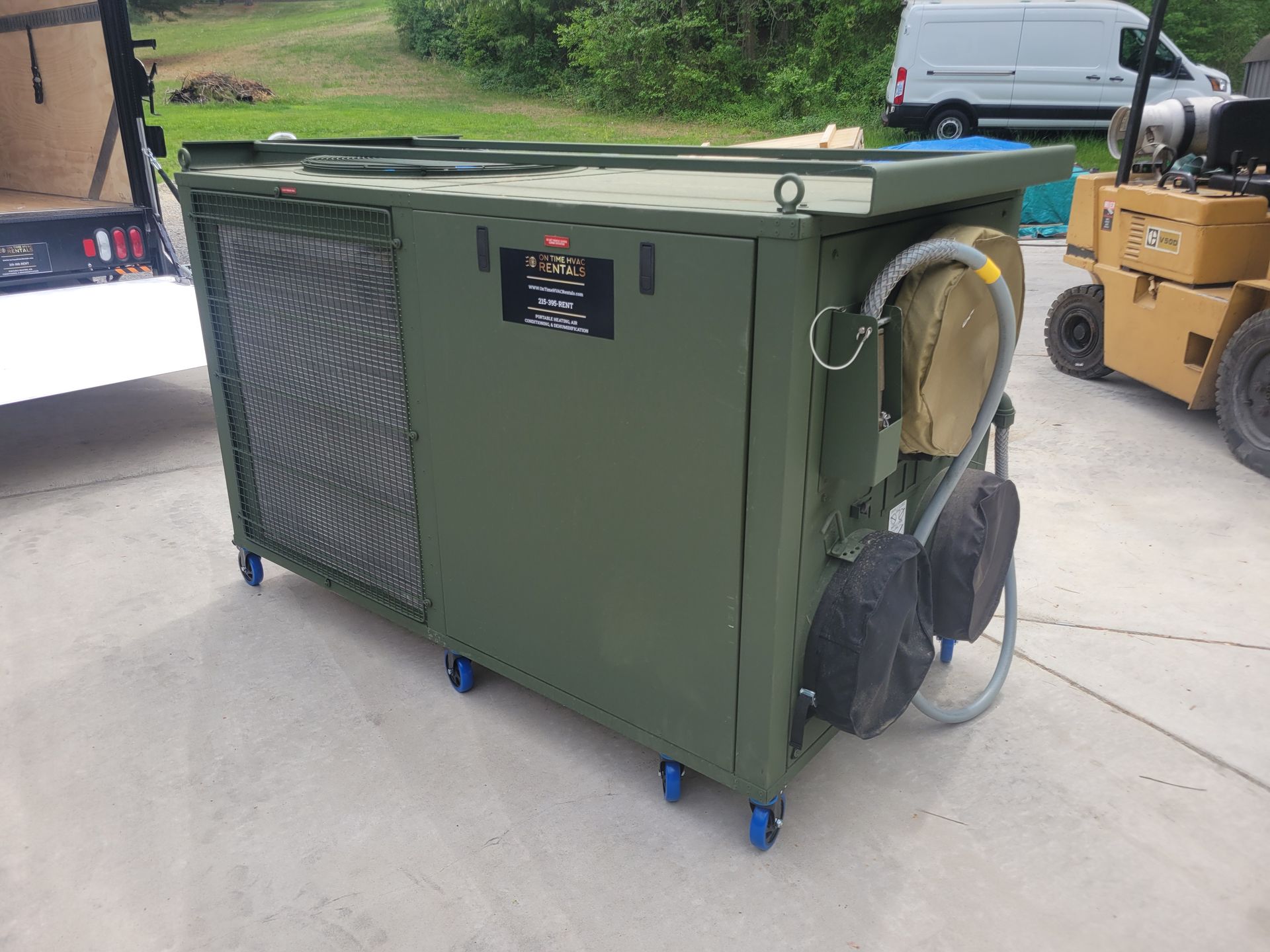 An olive-drab, military-style portable environmental control unit on wheels, parked on concrete with a forklift nearby.