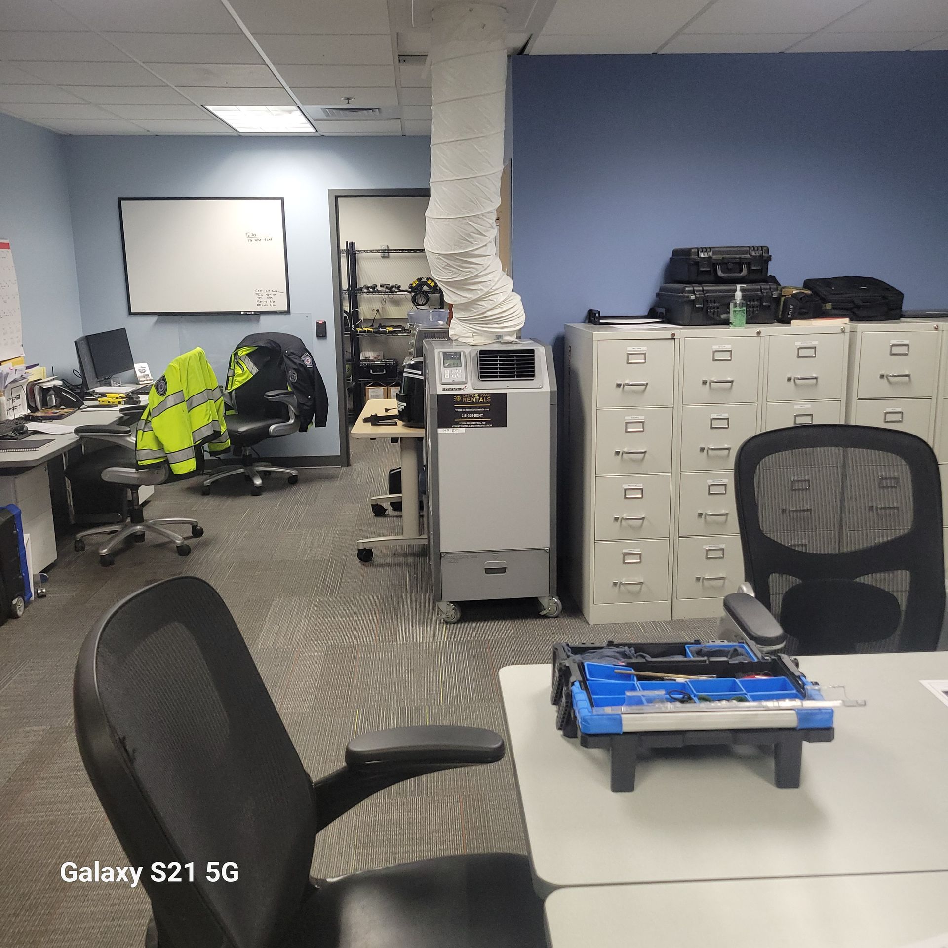 A cluttered office space featuring a portable air conditioner with an exhaust hose, rolling chairs, and filing cabinets.