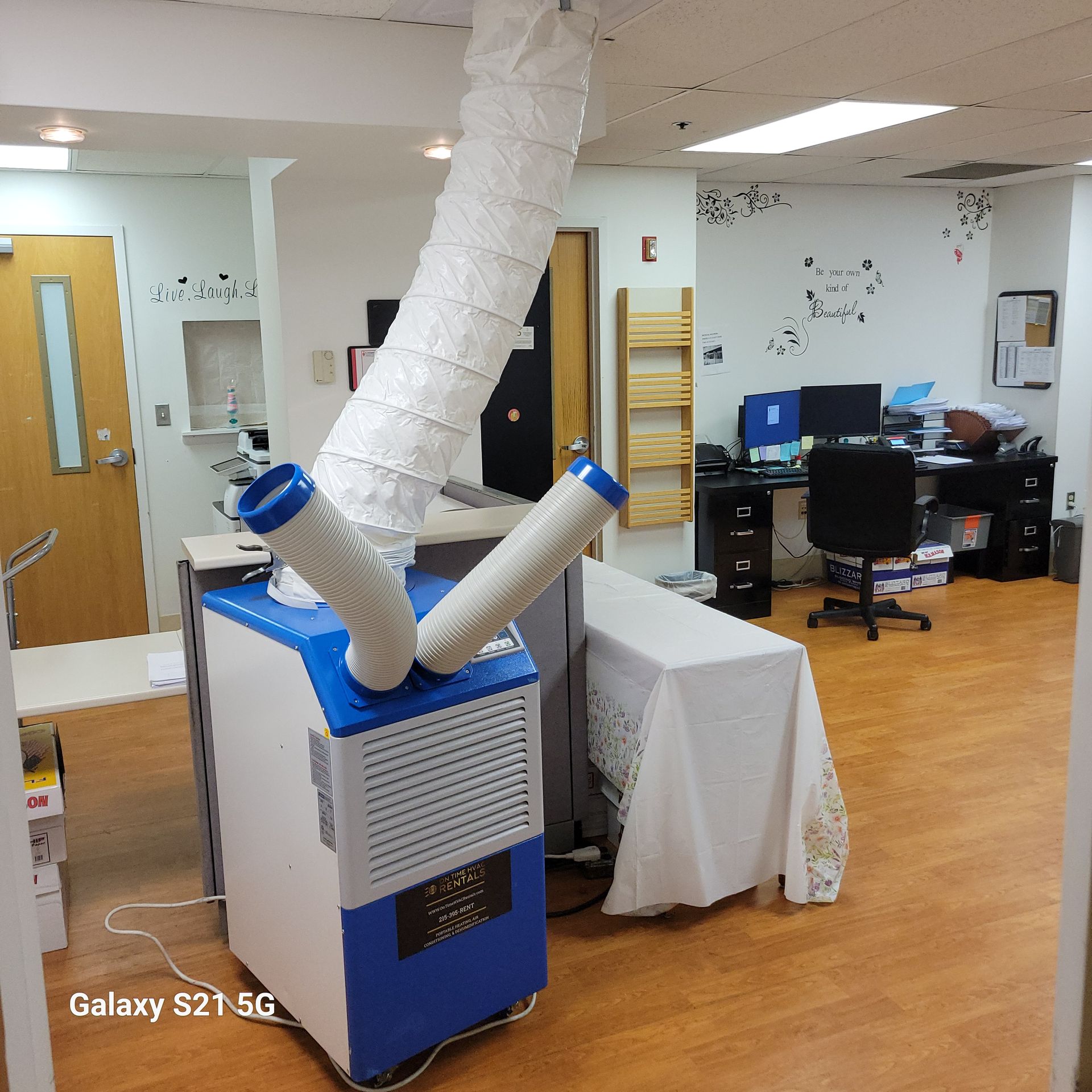 A portable dual-duct air conditioner stands on a wood floor in an office, venting through hoses toward the ceiling.