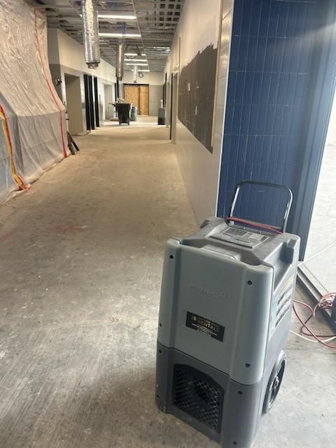 A commercial dehumidifier sits in a construction corridor with exposed ceilings, temporary wall plastic, and blue tiles.