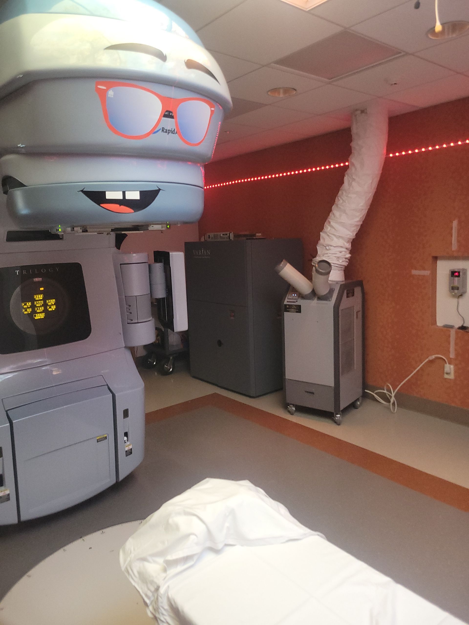 A medical radiation machine decorated as a friendly cartoon character with sunglasses, parked in a clinical treatment room.