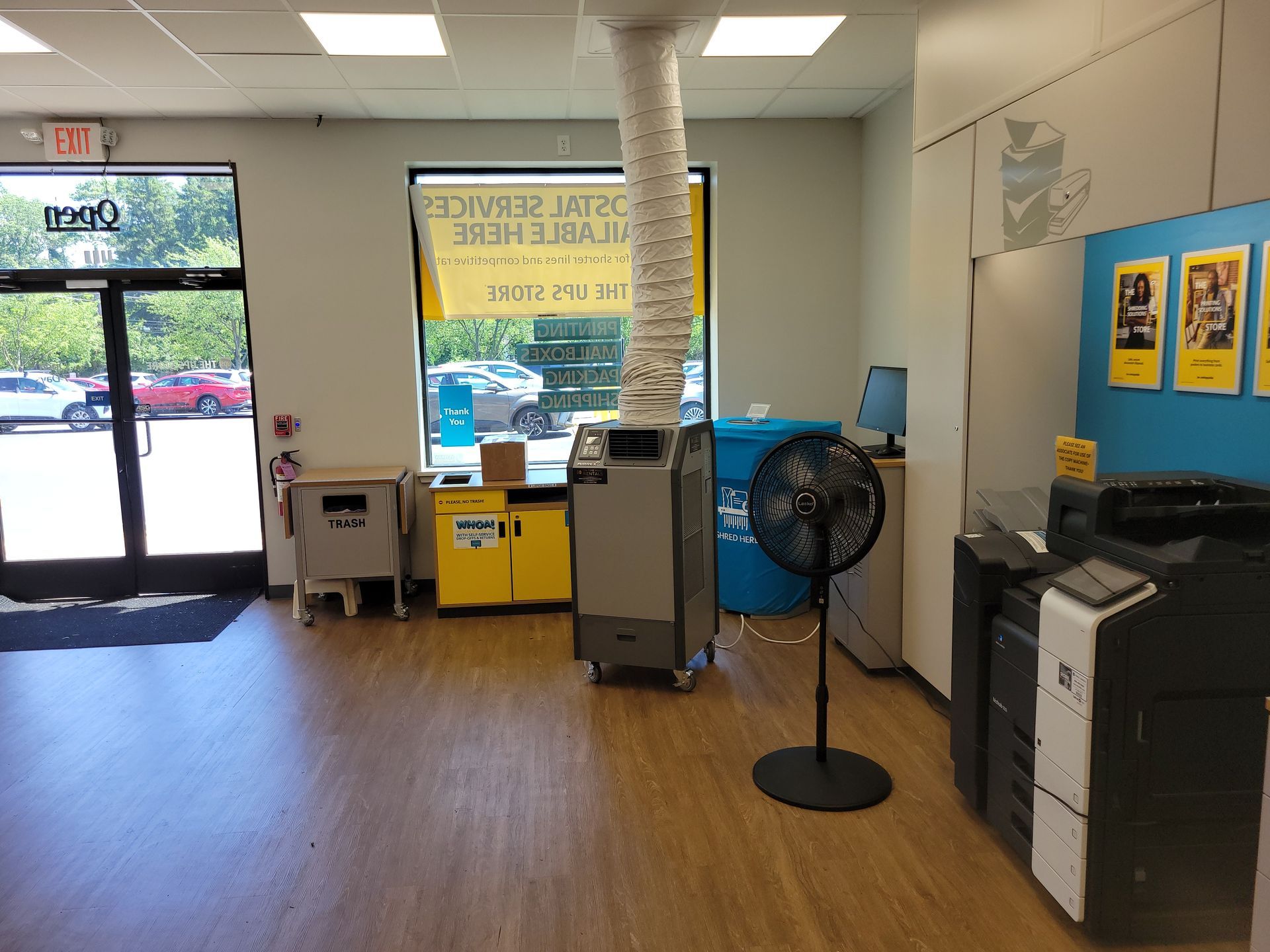 An office interior with a portable air conditioner connected to the ceiling, a standing fan, a copier, and store signage.