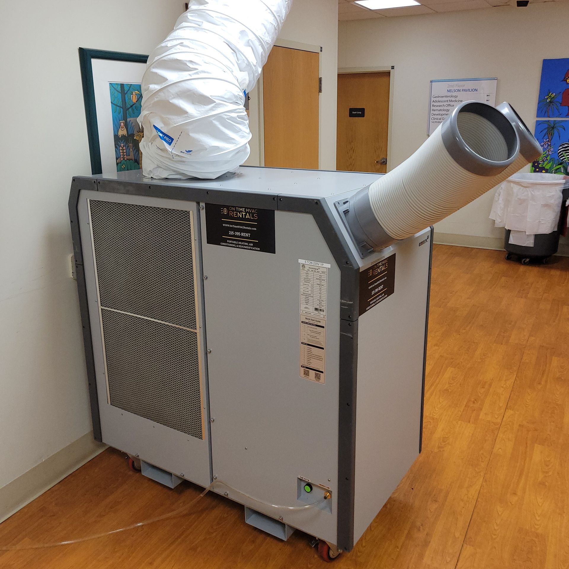 A portable spot air conditioner with flexible exhaust and cooling ducts on a wooden floor in a hallway.