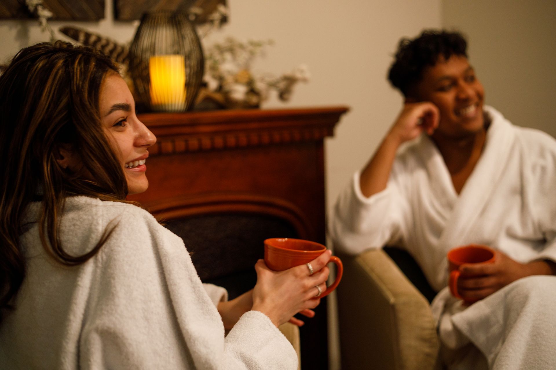 A man and woman relax in bathrobes near a fireplace, both holding coffee cups.