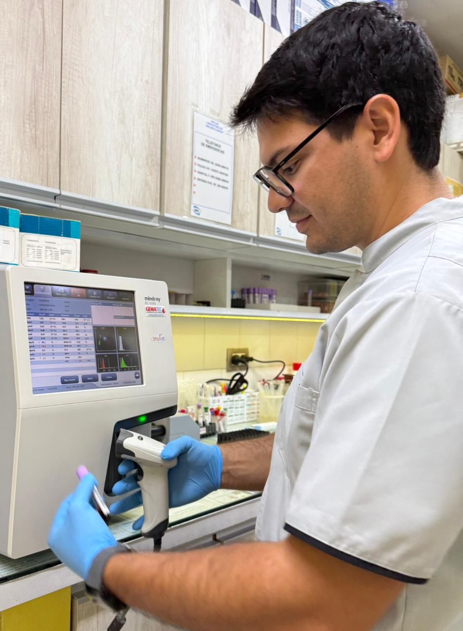 Un técnico de laboratorio con guantes azules usa una pipeta en una mesa junto a una máquina de laboratorio y un monitor.
