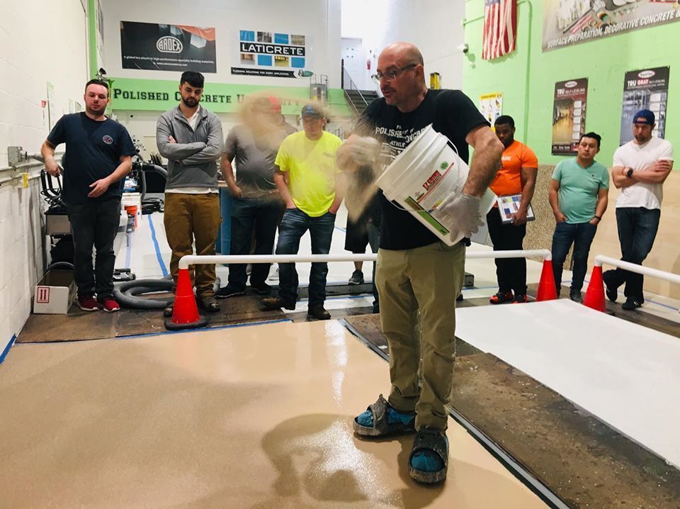 Man pouring powder over flooring, instructing a group. Indoors, concrete floor, several men observing.