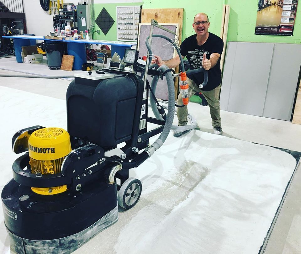 Man operating a large floor grinder, thumbs up, in a workshop. Dust and equipment present.